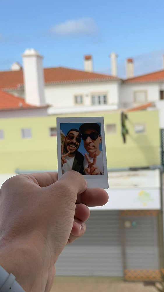A hand holds a polaroid photo of two friends smiling and posing together against a backdrop of buildings.