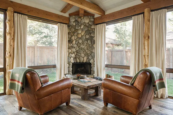 A rustic enclosed backyard sitting area with a central stone fireplace, exposed wood beams, and large windows wrapped in cream curtains. Two brown leather armchairs face a weathered wood coffee table on a plank floor, creating a cozy indoor-outdoor retreat.