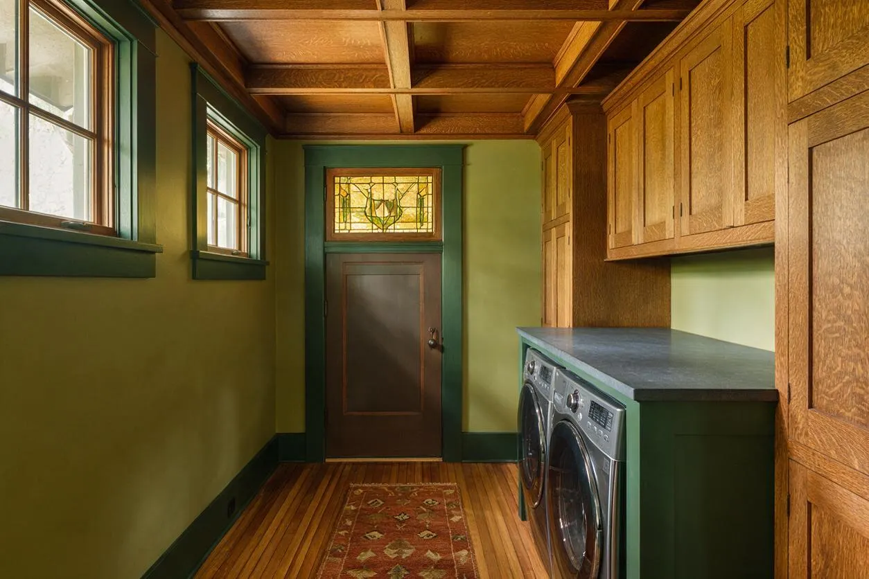 A narrow Craftsman-style laundry room with olive green walls, dark trim, and a coffered wood ceiling. Wood cabinets line one side above a front-loading washer and dryer, while a patterned runner, stained wood floor, and stained glass transom add warmth and character.