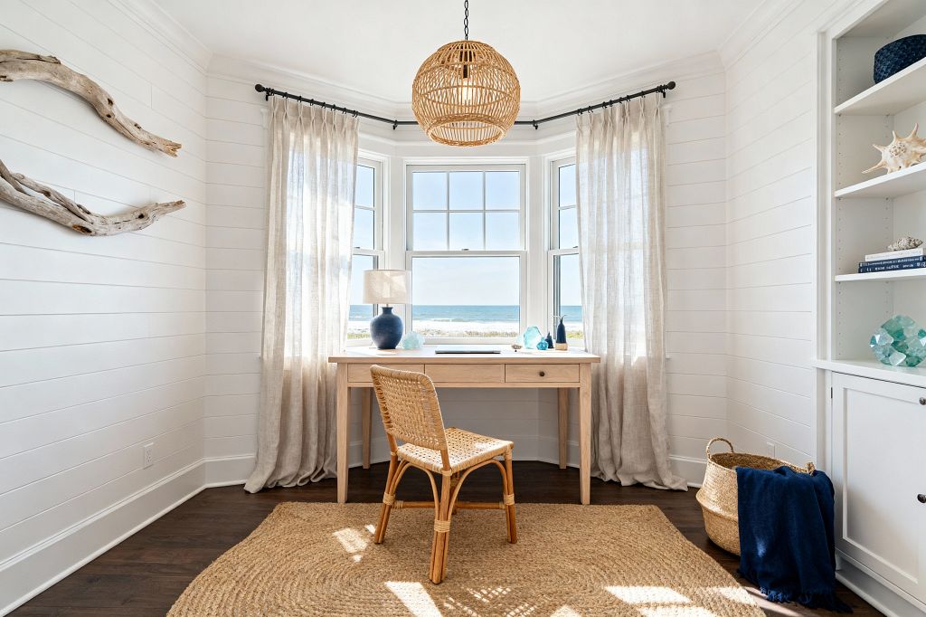 A bright coastal home office centers on a light wood desk set in a bay window overlooking the ocean. White shiplap walls, sheer beige linen curtains on a black rod, and a woven rattan pendant create an airy feel. A rattan chair sits on a round jute rug atop dark hardwood floors. Driftwood art hangs on the left wall, while built-in white shelves display sea-glass decor and shells. A blue ceramic lamp and a seagrass basket with a navy throw add color.