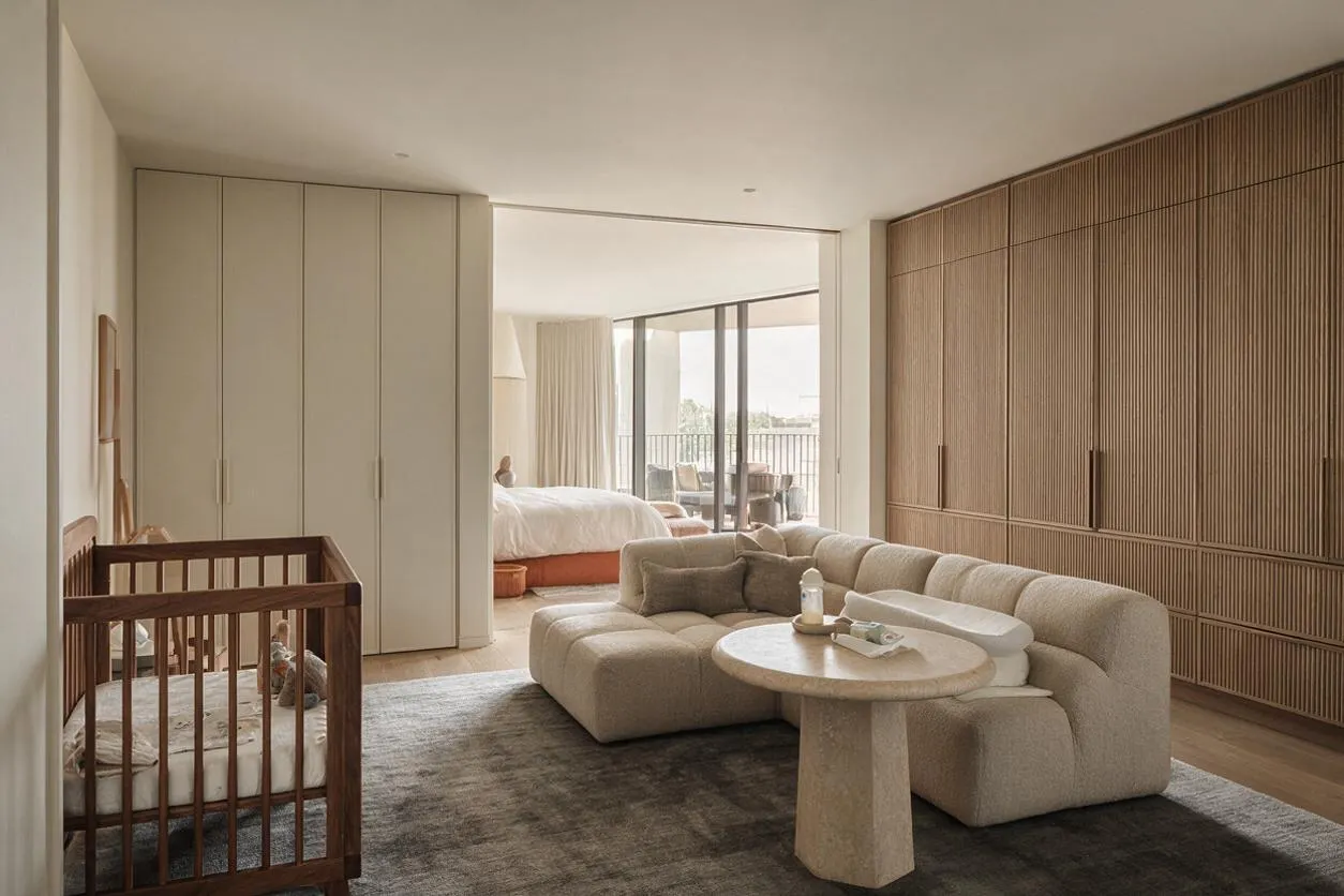 A modern nursery with a wooden crib on the left, a curved beige sectional sofa, and a round stone table on a dark rug. Full-height built-in wardrobes and a slatted wood feature wall frame the room, while a bedroom and balcony are visible through an open passage in the background.