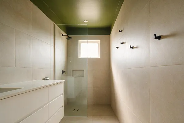 A modern master bathroom with large beige tiled walls and floors, a white floating vanity on the left, and a glass-enclosed shower at the back. The ceiling is painted olive green, and matte black shower and towel hooks add contrast to the clean, minimalist space.