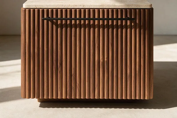 A modern cabinet with vertical fluted wood paneling in a warm brown finish and a light stone top. A slim black handle spans the upper front, and soft sunlight casts shadows across the textured surface.