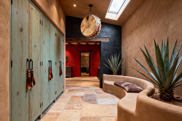 A Southwestern mudroom features tan, hand-troweled plaster walls and a curved built-in bench with agave planters and a brown cushion. The floor is multi-size tumbled stone tile in sand and terracotta tones. On the left, tall distressed turquoise wood lockers have black iron handles with leather strap pulls. Overhead, a rawhide drum pendant hangs near a skylight. A charcoal accent wall and timber beam frame a red stucco room beyond.