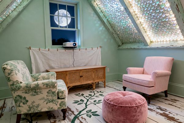 An attic media room with mint green walls, distressed rafters, and sloped skylights packed with iridescent glass discs that glow softly. A rattan cane cabinet holds a projector aimed at a linen sheet hung as a screen, facing two vintage armchairs—one in cream-and-sage toile, the other in pink ticking stripes—and a blush velvet pouf on worn white floorboards painted with a leafy vine; a night window frames a bright full moon.