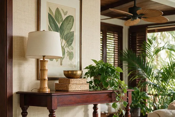 A tropical-style living room vignette features a dark wood console table topped with a bamboo table lamp, woven tray, brass bowl, and trailing green houseplant. Above it hangs framed botanical wall art, while louvered shutters, a palm plant, and a wood-blade ceiling fan add to the warm, airy atmosphere.