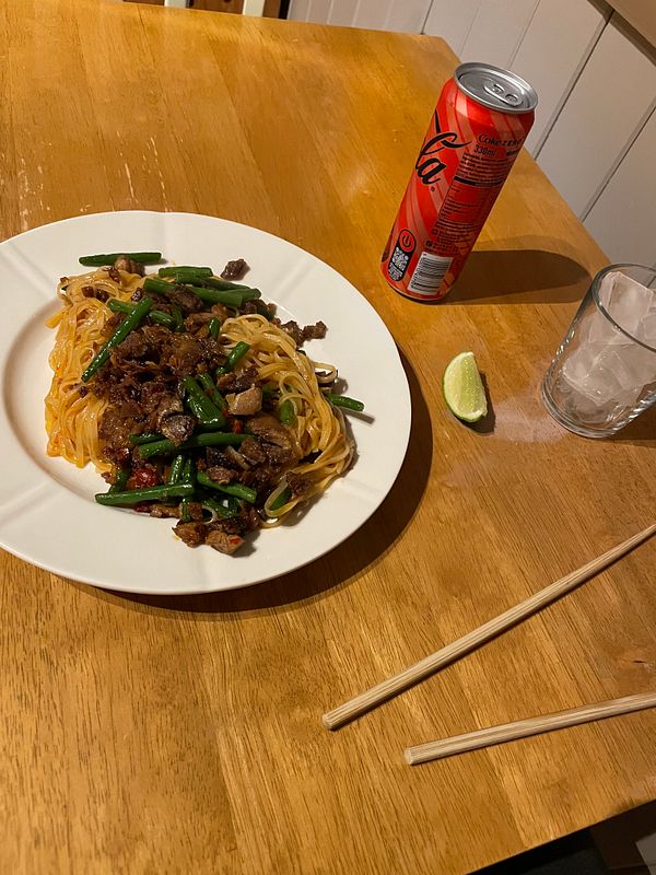 A plate of stir-fried noodles with pork belly and green beans is presented on a wooden table.