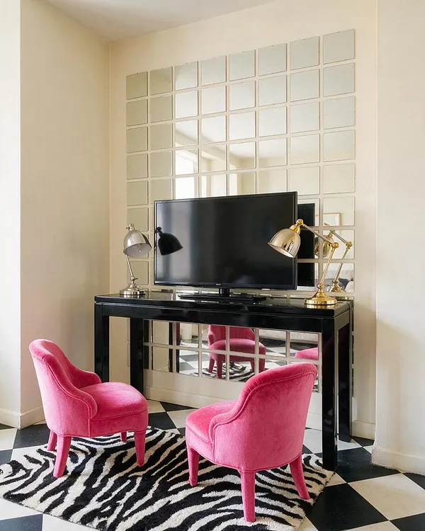 A small studio apartment corner styled in Hollywood Regency fashion, featuring a glossy black console with a flat-screen TV, two hot pink velvet chairs, and a large grid of mirrored tiles covering the wall behind it. A zebra-print rug sits over a black-and-white checkerboard floor, while metallic desk lamps add a polished, glamorous finish.