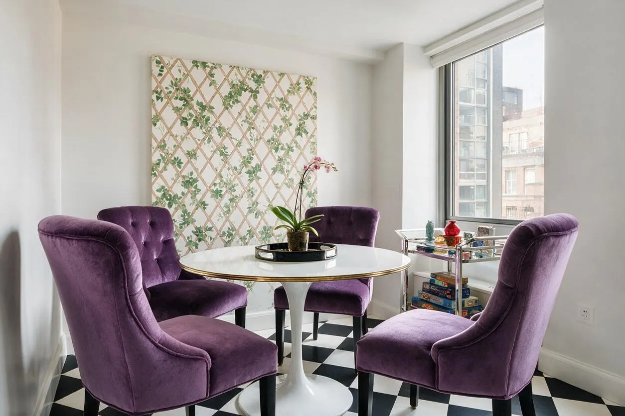A bright game room features four purple velvet dining chairs arranged around a round white pedestal table with a gold rim. The room has black-and-white checkered flooring, a floral lattice wall panel, and a window with a small acrylic and chrome bar cart beside it.