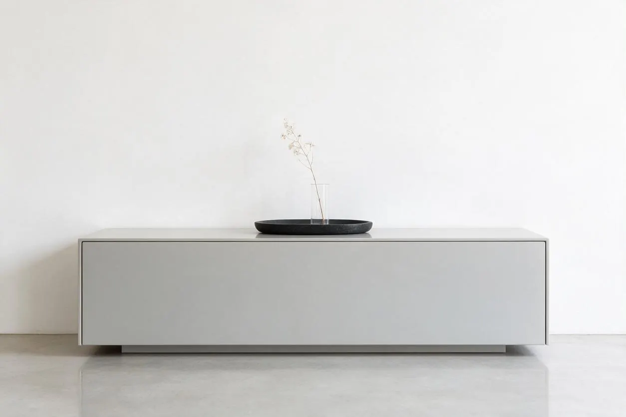 A sparse minimalist interior with a low, rectangular light gray cabinet centered against a plain white wall. On top sits a shallow black tray holding a clear glass vessel with a single delicate dried stem, creating a simple sculptural focal point.