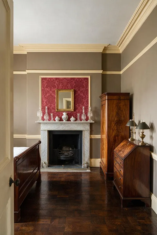 A traditional guest bedroom with taupe walls, cream crown molding, and dark hardwood floors. The room centers on a marble fireplace with a red damask panel above it, flanked by antique-style decor, a tall wood armoire, and a secretary desk with brass lamps.
