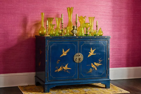A deep blue cabinet with gold bird and branch motifs stands against a bright pink textured wall. The cabinet top is styled with an eclectic row of yellow-green glass vases and candleholders, creating a bold maximalist vignette.