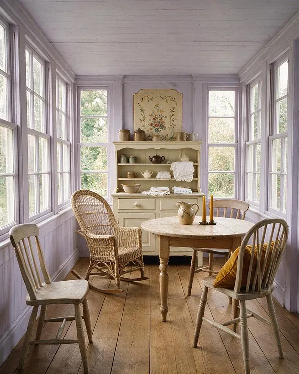 A narrow front porch styled as a cozy cottagecore dining nook, with lavender-painted walls, many tall windows, a round wooden table, mismatched wooden chairs, and a wicker rocking chair. A cream hutch with floral decor and pottery sits at the far end, while soft natural light warms the wood floor.