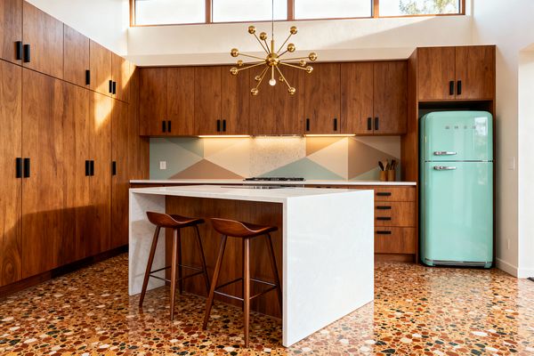 A Mid-Century Modern kitchen with full-height walnut cabinets and black pulls surrounds a white waterfall island with two curved walnut stools on a multicolored terrazzo floor. A mint retro-style refrigerator sits to the right. Under-cabinet lighting highlights a geometric backsplash in sage, tan, and cream behind a built-in cooktop. A brass sputnik chandelier hangs beneath clerestory windows, adding warm illumination to the clean, wood-forward space.