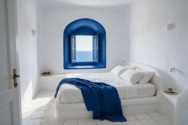 A minimalist Mediterranean-style master bedroom with whitewashed walls, a low white bed dressed in crisp white linens, and a vivid blue throw draped over the foot of the bed. A deep blue arched window frames a view of the sea, filling the room with bright natural light.