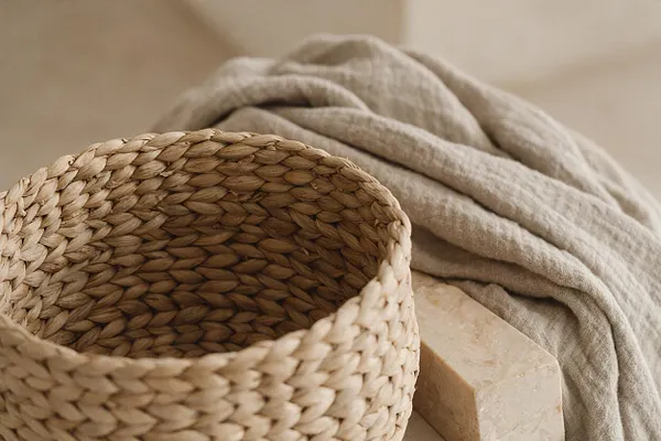 A close-up still life shows a woven natural basket beside a softly draped beige linen fabric and a light stone block. The scene uses warm neutral tones and tactile organic textures to create a calm Organic Modern look.