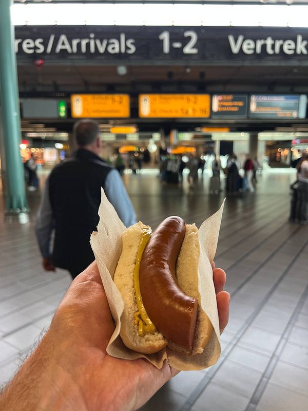 A hand holds a hot dog in an airport terminal with arrival and departure signs in the background.