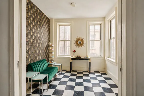 A bright sunroom with black-and-white checkerboard flooring, cream walls, and a dramatic Art Deco accent wall in gold and black fan pattern wallpaper. A green velvet sofa sits beside mirrored side tables, while a sunburst mirror and slender console add vintage glamour under tall windows.
