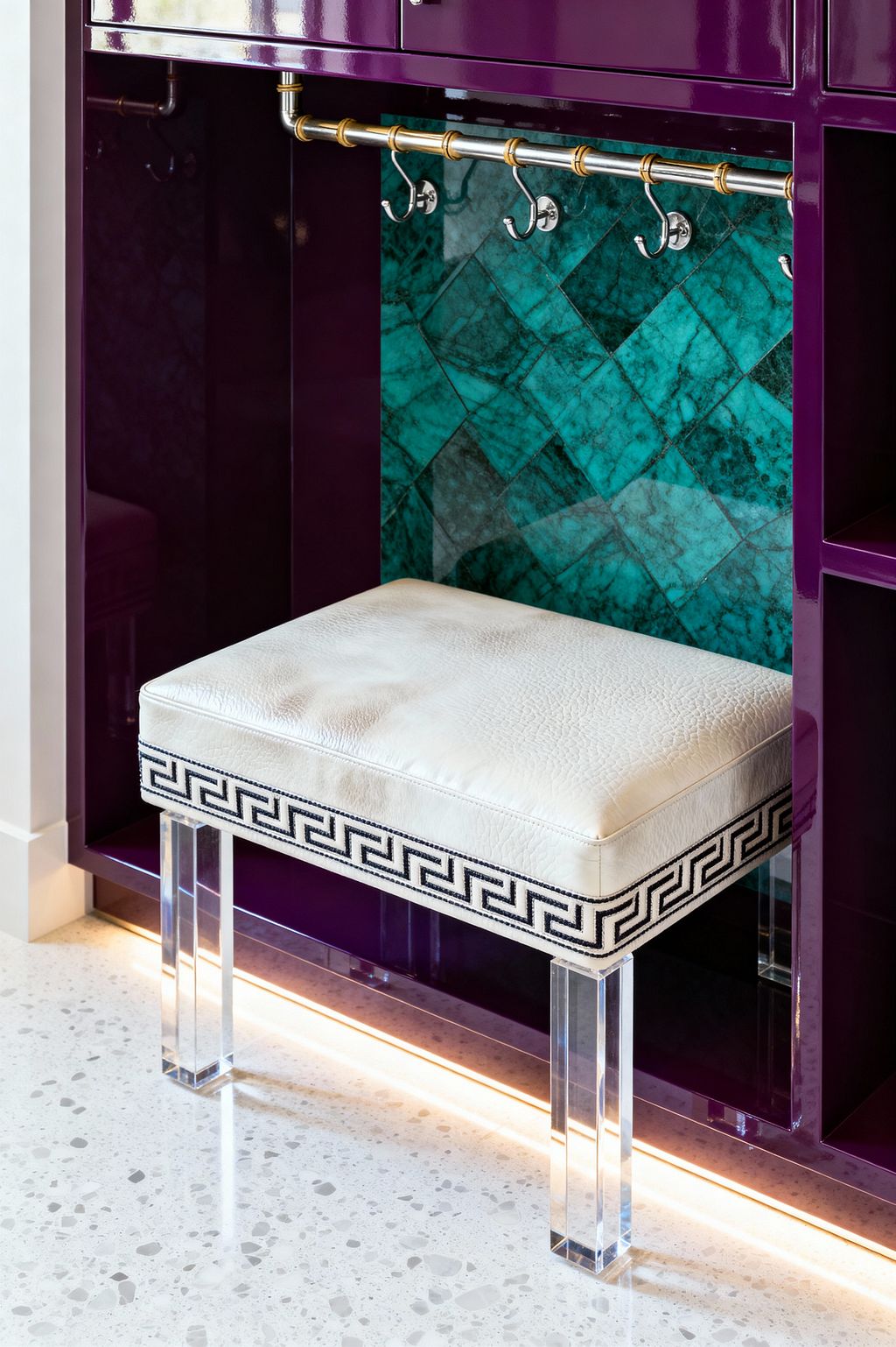 A mudroom nook features glossy plum lacquer cabinetry framing a bench with a white leather cushion edged in black Greek-key trim and supported by clear acrylic legs. Behind a brass-accented hanging rail with polished nickel hooks, emerald herringbone stone tiles create a jewel-toned backdrop, while warm LED toe-kick lighting highlights the light gray terrazzo floor with small aggregate flecks.
