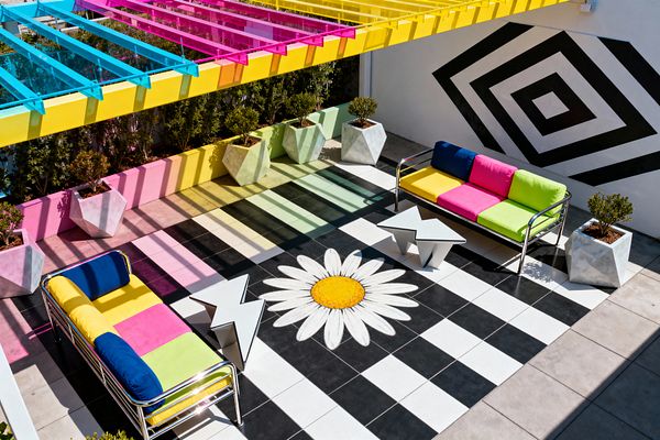 A vibrant outdoor patio features a CMY-tinted pergola that casts colored stripes onto a black-and-white checkerboard floor with a large daisy graphic at its center. Two chrome-framed sofas with cushions in cobalt blue, bright yellow, hot pink, and lime flank white zigzag side tables; a black concentric-diamond mural animates the white wall, and faceted concrete planters with shrubs border the space.