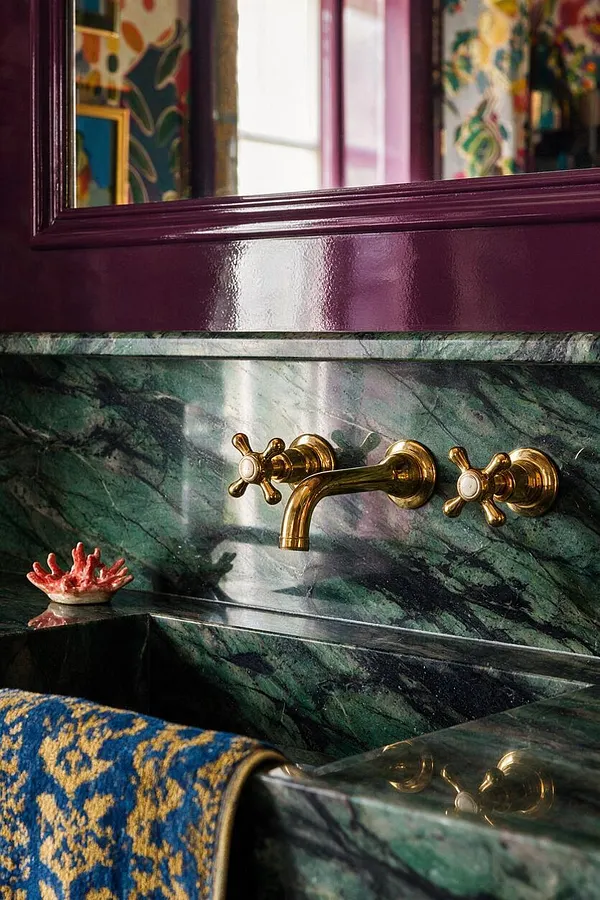 A close-up of a dramatic bathroom sink area with deep green marble or stone, glossy plum trim, and polished brass faucet handles and spout. A patterned blue-and-gold textile hangs over the foreground edge, adding to the richly layered maximalist look.
