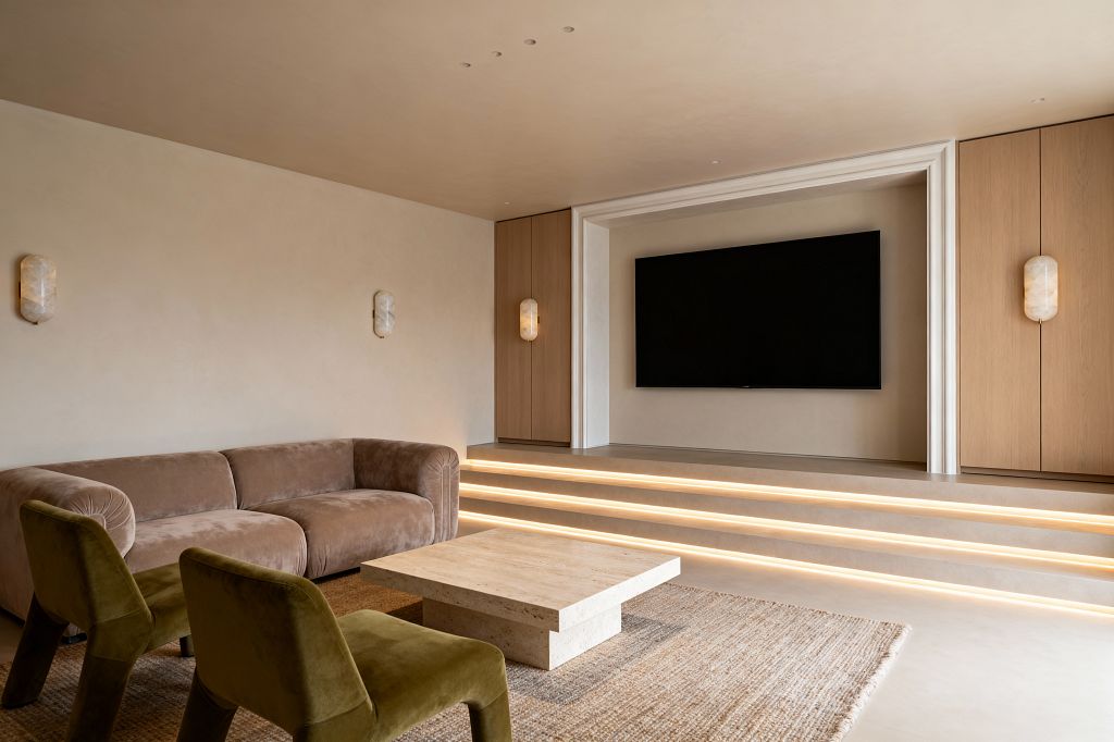 Neutral media room with a large wall-mounted TV set into a recessed niche, flanked by light oak panels and stone-like wall sconces. A taupe velvet sofa and two moss green upholstered chairs face a square travertine coffee table on a chunky jute rug, while tiered steps with integrated linear LED lights create a soft glow; smooth beige plaster surfaces and discreet recessed downlights convey a calm, transitional aesthetic.