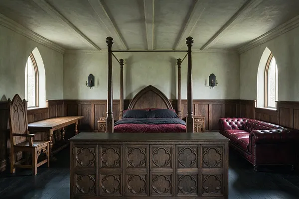 A Gothic-style bedroom with a carved four-poster bed centered against the back wall, flanked by small wooden nightstands and dark wall sconces. The room includes a red leather Chesterfield sofa on one side, a carved wooden chair and desk on the other, arched windows, paneled wood walls, and a dark floor under a pale vaulted ceiling.