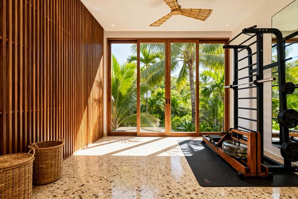 A bright home gym with floor-to-ceiling wood-framed sliding glass doors overlooks dense palm trees. Inside, a vertical slat hardwood wall lines one side, while a black steel training rack, wall mirror, and wood water rower sit on a rubber mat opposite. The floor is speckled terrazzo, and a woven rattan ceiling fan hangs above. Soft white walls and abundant natural light create a calm, tropical atmosphere, with two woven baskets in the corner.