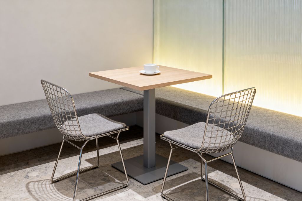 A minimalist dining nook features a square light wood table on a gray pedestal, flanked by a built-in gray felt bench and two chrome wire chairs with matching gray seat pads. The floor is neutral terrazzo, an off-white wall sits to the left, and a fluted glass partition behind the bench glows with warm concealed lighting, while a single white cup on the table underscores the uncluttered aesthetic.