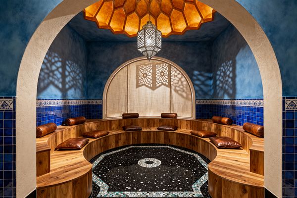 An intimate Moroccan-style media room is framed by a cream plaster arch and finished with textured blue walls and cobalt tile wainscoting. Curved tiered wood benches with brown leather cushions surround a central black terrazzo mosaic, while a warm amber honeycomb ceiling and a pierced metal pendant lantern cast patterned shadows onto an arched niche draped with a neutral curtain.