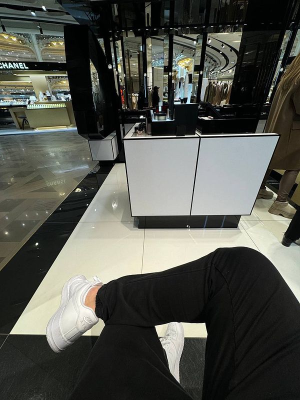 A person is seated in a Chanel store, showcasing their white sneakers and black pants.