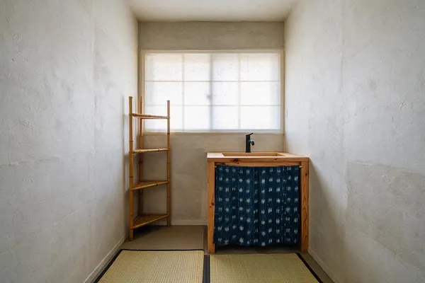 A narrow powder room designed in Japanese Zen style with textured off-white plaster walls, a small frosted window, and a simple natural wood vanity with a dark faucet and indigo fabric panel. A bamboo shelving unit stands to the left, and woven tatami mats cover part of the floor.