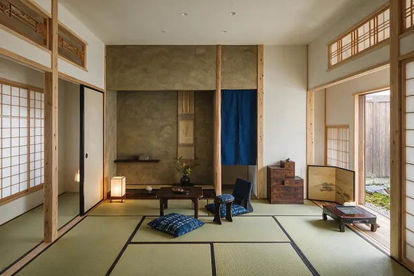 A serene Japanese Zen-style front porch or entry room with tatami mat flooring, exposed light wood posts, shoji sliding screens, and an alcove with a hanging scroll. Low seating, blue cushions, a lantern, and simple wood furnishings create a calm, minimalist atmosphere with natural light.