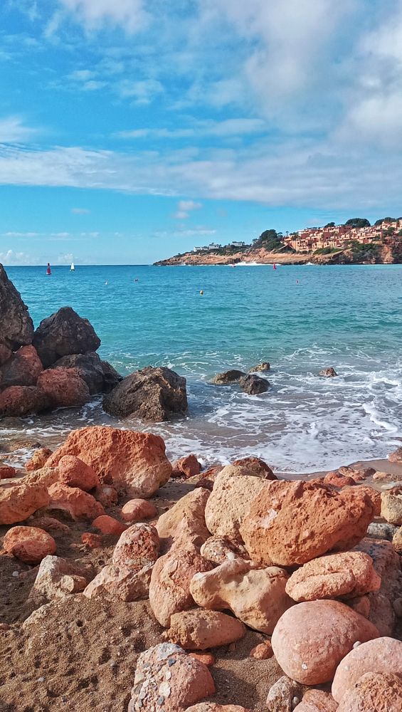 A serene beach scene showing a sandy shore, blue sea, a small rocky island, and a distant mountain under a partly cloudy sky.