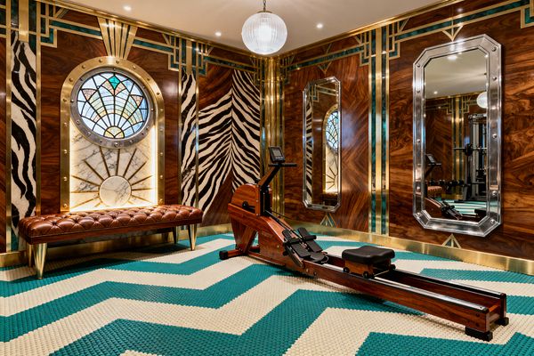 Art Deco home gym with bookmatched walnut wall panels trimmed in brass and emerald accents, featuring zebra-patterned sections. A round stained-glass window set in a marble arch with a brass sunburst glows above a tufted cognac leather bench. Octagonal chrome-framed mirrors line the opposite wall. The floor shows teal and ivory chevron rubber coin tiles, and a wooden water rower sits centered under a ribbed glass globe pendant and recessed ceiling lights.