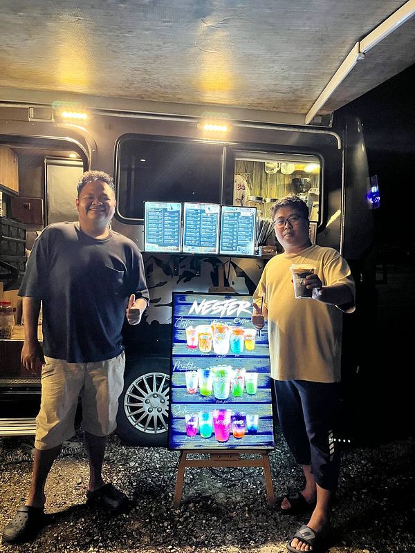 Two men stand in front of a coffee motorhome, showcasing a colorful drink menu.