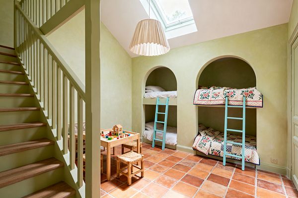 A children's room in French Country style with sage green limewashed walls features two built-in arched alcoves housing stacked bunks. Patchwork floral quilts and white bedding sit behind pale turquoise ladders. Rustic terracotta floor tiles, a natural wood play table with woven rush-seat stools, a green-painted staircase, a skylight, and a pleated fabric pendant complete the warm, sunlit space.