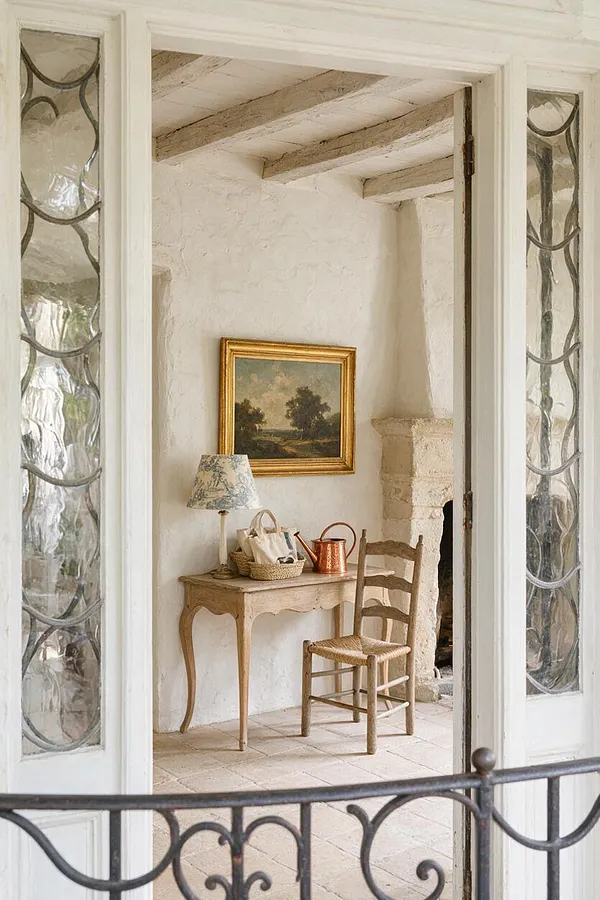 A French Country front porch with white plaster walls, exposed ceiling beams, and a stone fireplace. A small antique wooden table holds a floral lamp, baskets, and a copper watering can, with a woven chair placed beside it. Leaded glass doors and wrought iron railing frame the cozy entry.
