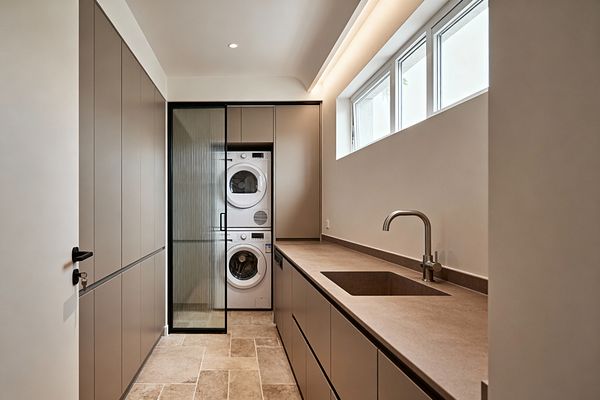 A narrow, minimalist laundry room with warm greige, handleless cabinets lines both sides. A long stone-look countertop with an integrated sink and brushed metal faucet runs beneath clerestory windows and a linear cove light. At the far end, a black-framed sliding door of reeded glass reveals a stacked white washer and dryer built into matching cabinetry. Beige stone-look floor tiles complete the calm, uncluttered space.
