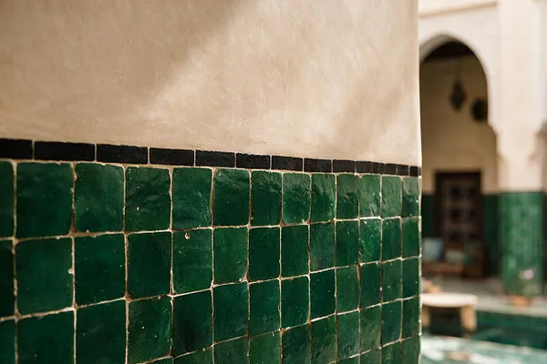 A Moroccan-style interior wall features glossy deep green square zellige tiles set beneath a smooth beige plaster surface and a thin black border. In the softly blurred background, an arched opening and more green tilework suggest a traditional courtyard or riad.