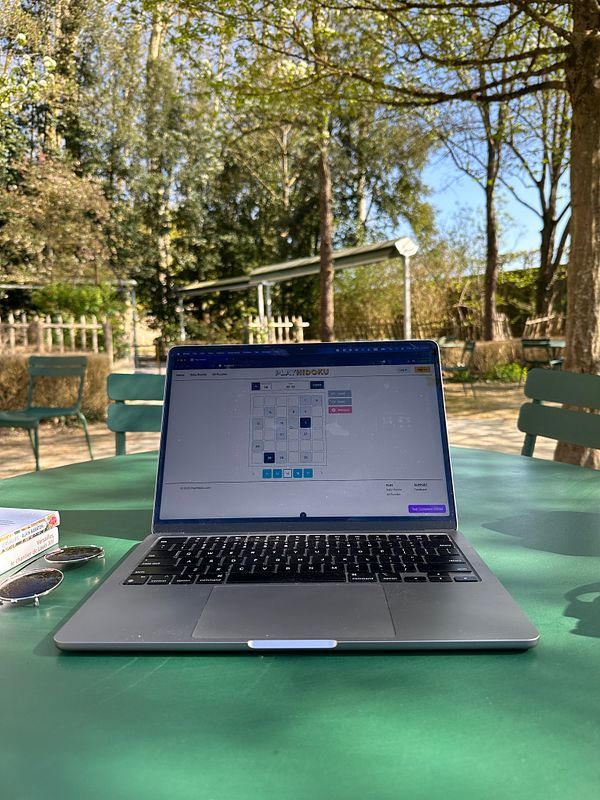 A laptop is set up on a green table in a serene outdoor setting, displaying a Sudoku game interface.