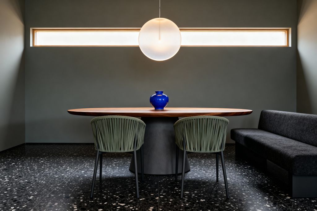 A modern dining room features an oval walnut table on a gray pedestal, flanked by two sage green rope-back chairs and a charcoal upholstered bench. A large frosted globe pendant hangs centered before a narrow horizontal clerestory window. The room has matte gray-green walls and black terrazzo flooring with white flecks; a bright cobalt vase sits on the tabletop.