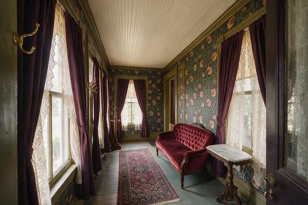 A narrow Victorian front porch lined with tall windows dressed in plum velvet drapes and lace sheers. The space features floral wallpaper, a tufted burgundy loveseat, a small marble-topped side table, a patterned runner rug, and brass wall hooks, creating a richly layered period look.