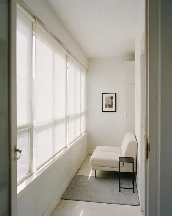 A narrow minimalist sunroom with large floor-to-ceiling windows on the left covered by sheer white shades, filling the space with diffused light. The room features a small white chaise lounge, a black side table, a light gray rug, and a framed black-and-white artwork on the back wall, all set against crisp white walls.