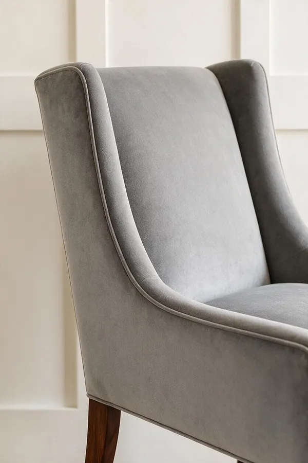 A close-up of a transitional upholstered chair in soft gray velvet, showing a gently curved wingback silhouette with tailored piping and a dark wood leg. The chair sits against a white paneled wall, creating a refined, calm backdrop.