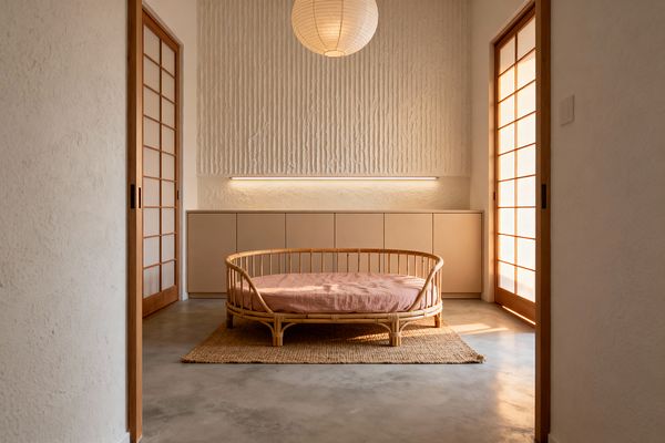 A minimalist kids’ room features a low circular rattan daybed with blush linen on a jute rug, centered on a polished concrete floor. Warm white plaster walls include a vertically ribbed accent and a long built-in cabinet with beige fronts, lit by a slim linear LED; wood-framed shoji doors filter sunlight from both sides, and a round rice-paper lantern hangs above.
