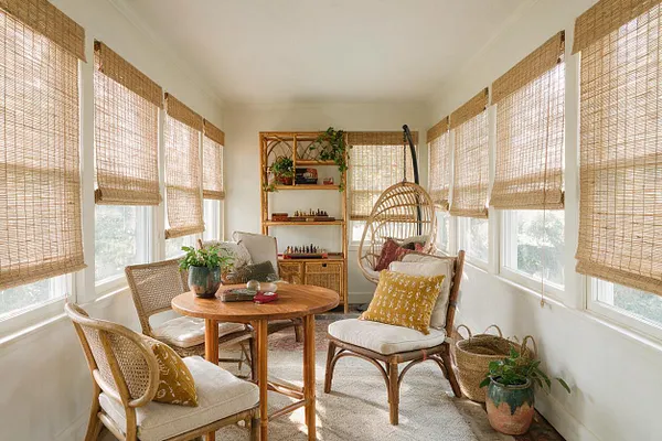 A bright bohemian game room with woven bamboo blinds on all the windows, a round wood table, and several rattan and cane chairs with cream cushions. A hanging egg chair, open shelving with decor and game pieces, potted plants, and a light neutral rug create a relaxed, natural atmosphere.