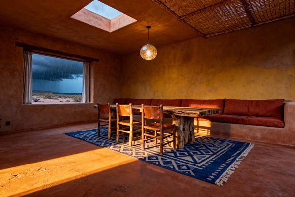 A Southwestern living room with earthen plaster walls and a terracotta floor features a built-in corner bench with rust-colored cushions, a live-edge wood table, and four leather sling chairs set on a blue-and-ivory geometric wool rug. A large picture window with simple white curtains frames a rainy desert view, while a square skylight, a globe pendant, and a woven reed ceiling panel cast warm light and texture.