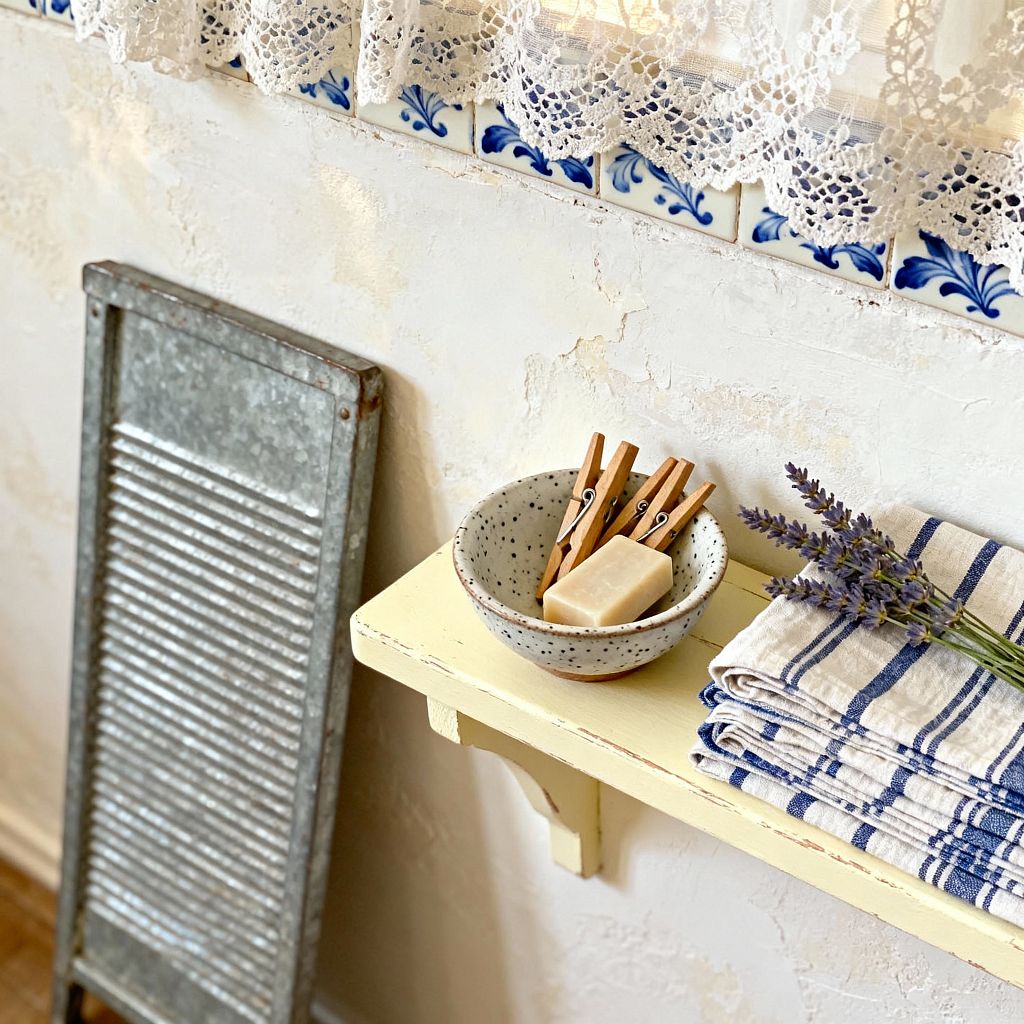 A farmhouse laundry vignette features a small butter-yellow wall shelf with a speckled stoneware bowl holding wooden clothespins and a natural soap bar, next to folded blue-striped towels and a sprig of lavender. Behind, a textured cream plaster wall is trimmed with blue-and-white floral tiles and a scalloped lace curtain, while a galvanized metal washboard leans nearby, all illuminated by gentle daylight that highlights the aged, handcrafted surfaces.