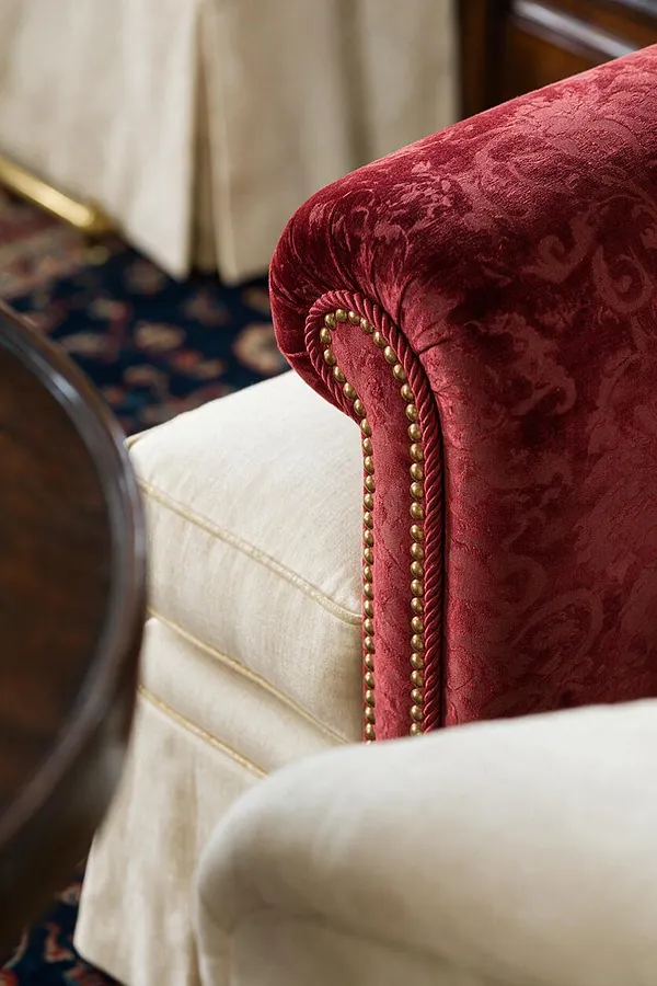 A close-up of a traditional upholstered sofa arm in rich crimson velvet with a subtle damask pattern, trimmed with twisted cord and brass nailhead detailing. Cream upholstered seating and a dark patterned rug appear softly in the background, along with warm wood and brass accents.